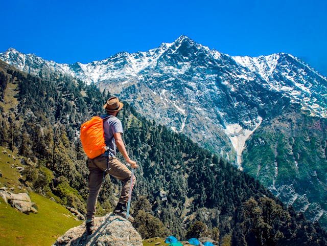 A hiker wearing a hat and carrying an orange backpack stands on a rock, gazing at a towering snow-capped mountain range under a clear blue sky. The surrounding landscape is covered in lush green trees and grassy slopes, with a few tents visible in the distance.