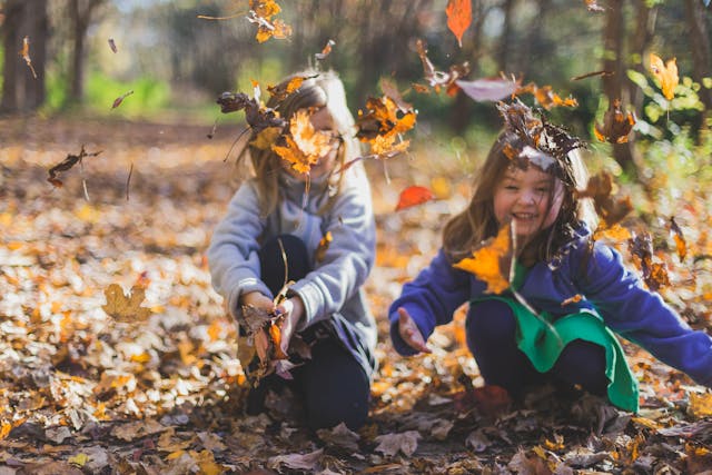 Two young girls playfully throw autumn leaves in the air while crouching on a forest path covered in fallen leaves. One girl wears a light-colored sweater, while the other wears a bright blue coat with a green dress. They both have joyful expressions as they enjoy the crisp fall day.