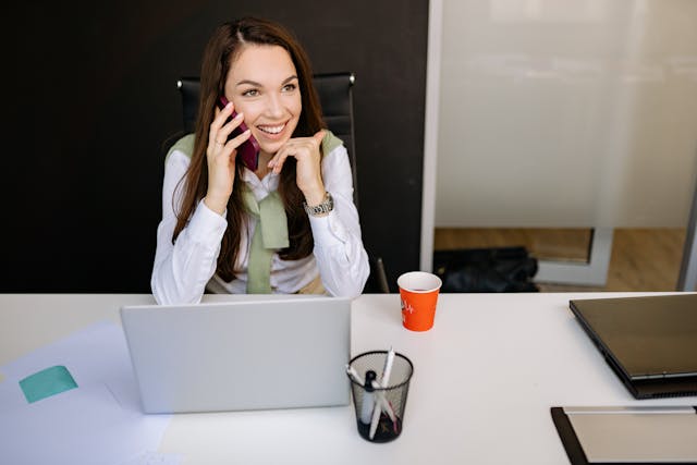 woman-smiling-while-talking-on-the-phone-at-her-desk-with-a-laptop-papers-and-a-coffee-cup-in-front-of-her